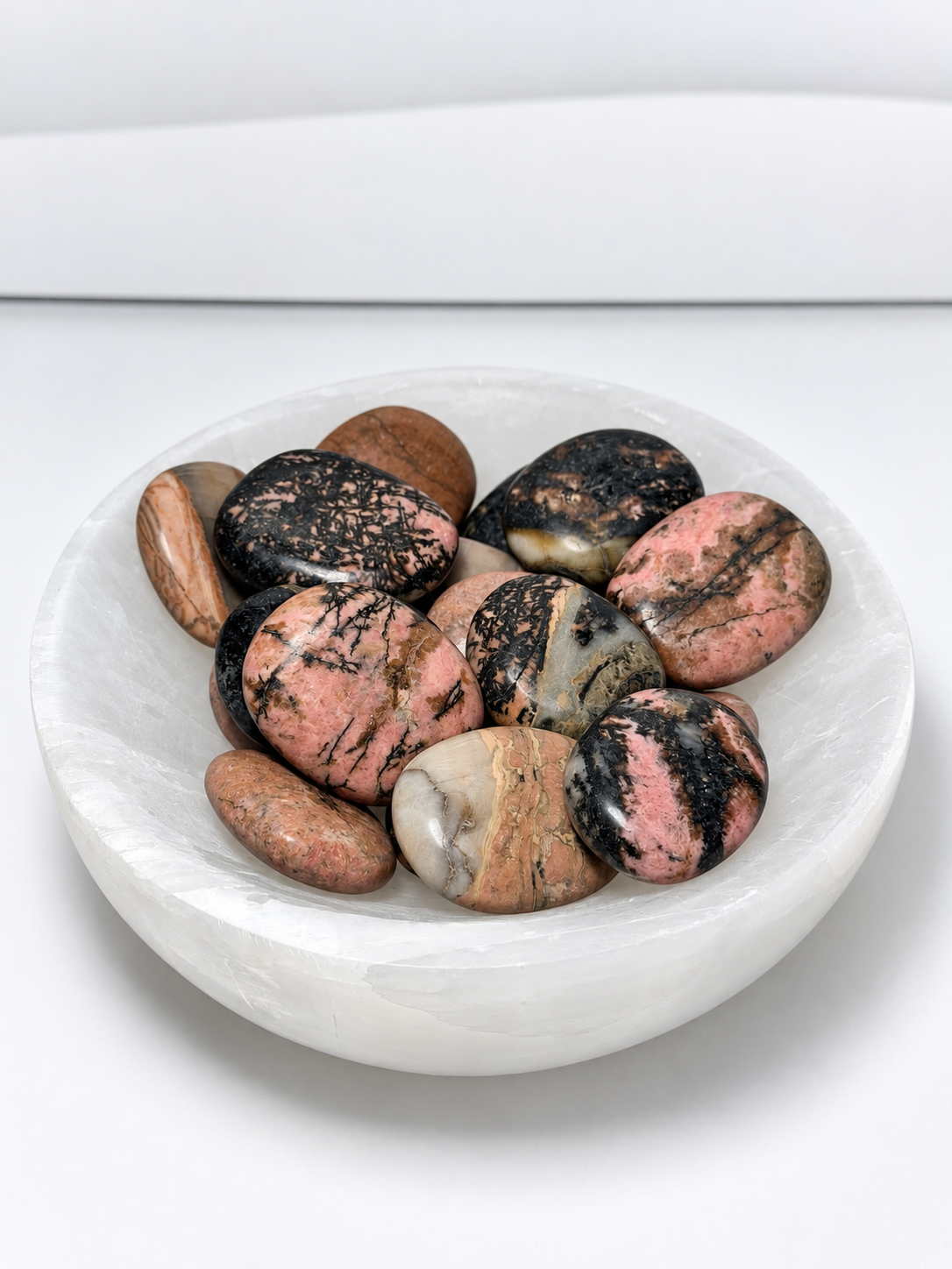 Rhodochrosite polished palmstones in pink, black, cream, and earthy tones displayed in a white crystal bowl